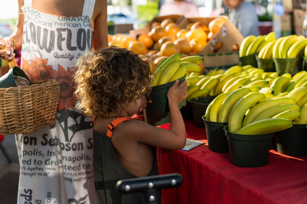 Young child reaching for fresh bananas and oranges at a market stand